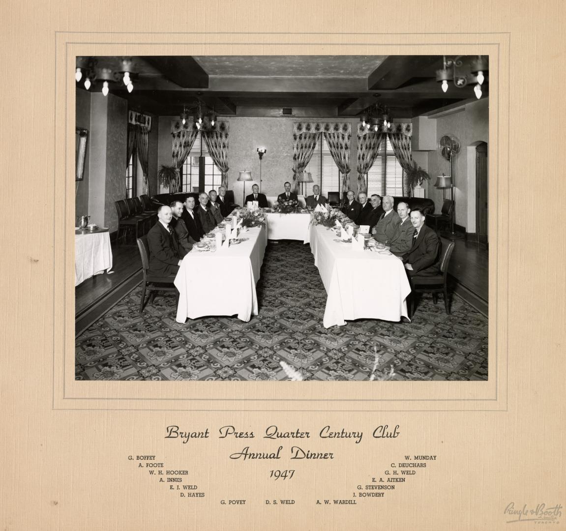 A photograph of a group of people posing around a long banquet table, which is set with dinnerw…