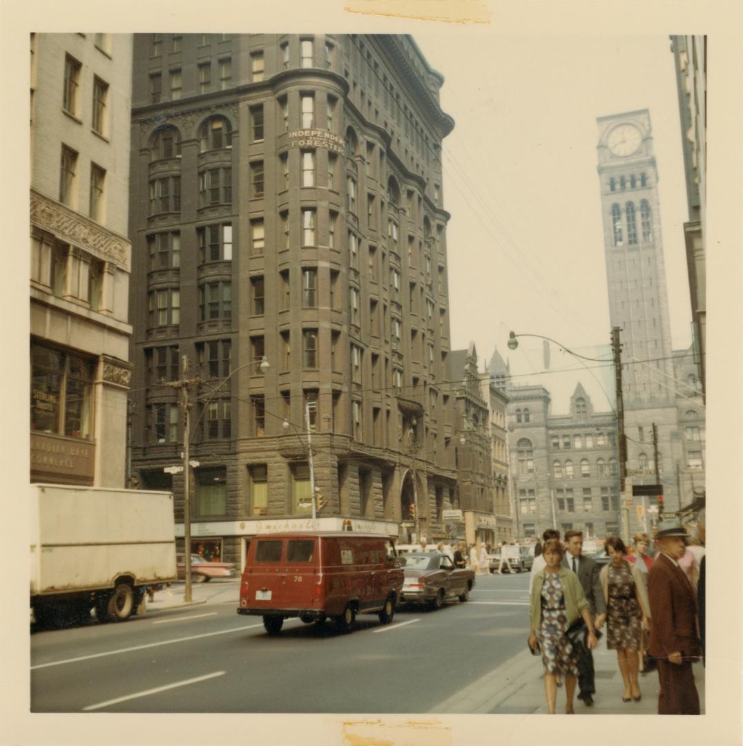 A photograph of a city street, with tall stone buildings on the left side of the photo and a bu…