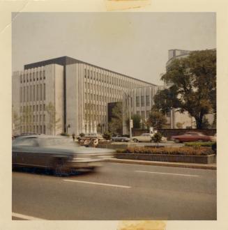 A photograph of a large brutalist building beside a wide, paved city street. There is a large t…