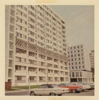 A photograph of a thirteen story apartment building, with a paved city street in front of it an…