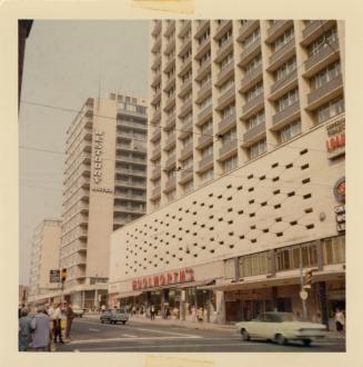 A photograph of a downtown city street, with a paved road in front of a line of skyscrapers and ...