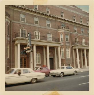 A photograph of a four story building, with a paved city street and sidewalk in front of it. Th…