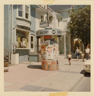 A photograph of a paved city sidewalk, with a row of houses or businesses on the left side and …