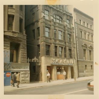 A photograph of a row of four and five story stone or brick buildings. The one in the middle of…