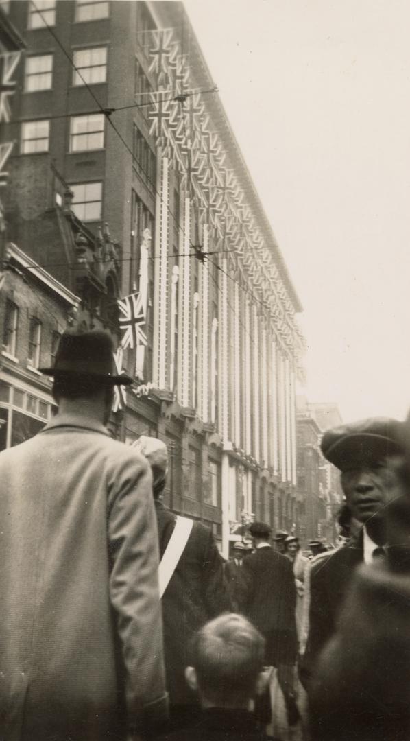 A photograph of a tall building facing a city street, decorating with bunting and numerous flag…