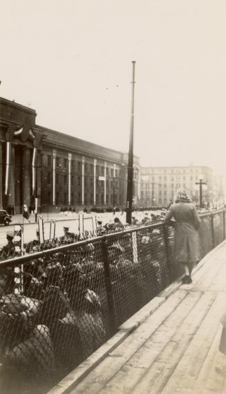 A photograph of a crowd of people standing across a road from a large building with stone colum…