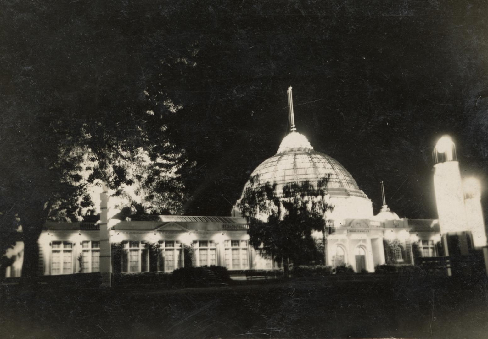 A photograph of a building with a glass dome, lit up at night. There are trees in front of the …