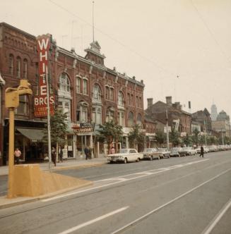 A photograph of a row of brick buildings beside a paved city street. There are cars parked on t…