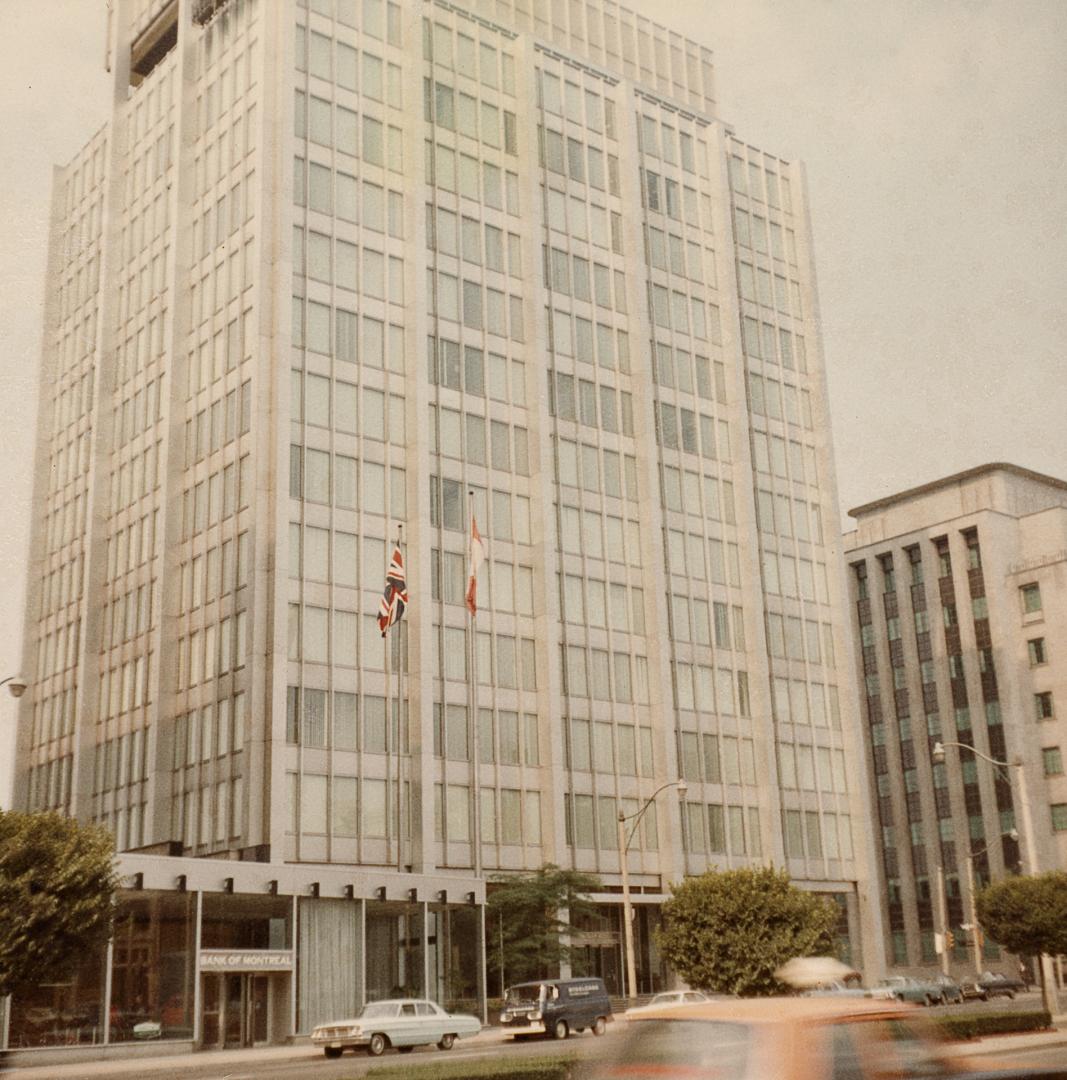 A photograph of two office buildings beside a paved city street. There are cars parked on and d…