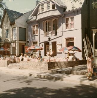 A photograph of a row of townhouse-style buildings beside a paved city street. There is a patio…