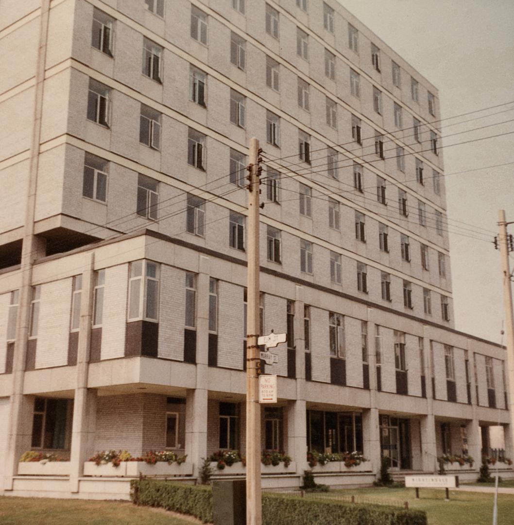 A photograph of a concrete eight story office building, with a grass lawn in front of it and ut…