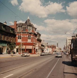 A photograph of a paved city street, with townhouse-style buildings on both sides of the street…