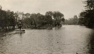 A photograph of a body of water separating wooded areas. There is a five-tiered fountain on the…