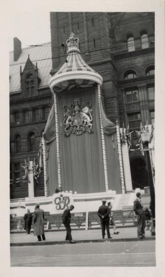 A photograph of a stage erected in front of a large brick building. The stand is decorated with…