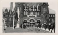 A photograph of a large stone building decorated with bunting, banners and flags. There is a pa…