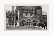 A photograph of a large stone building decorated with bunting, banners and flags. There is a pa…