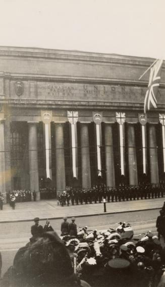 A photograph of a large stone building with columns decorated with banners and crests. There is…