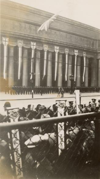 A photograph of a large stone building with columns decorated with banners and crests. There is…