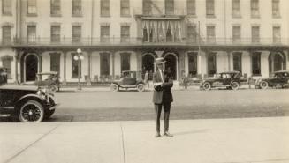 A photograph of a person standing on a sidewalk, with a hotel behind him on the other side of a…