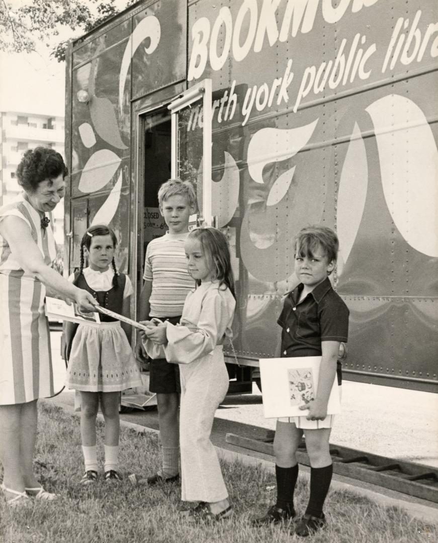 Picture of female librarian, standing in front of a bookmobile, handing a books to a child with…