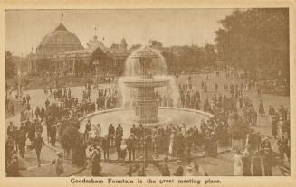 Sepia toned photograph of crowds of people standing around a huge fountain. Large buildings are…