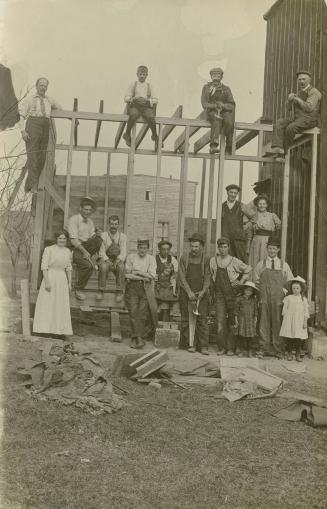 Black and white photograph of men, women and children posing in front of the frame of a house u…