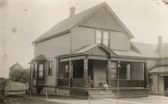 Black and white photograph of a of a small girl sitting on the steps of a two story frame house…