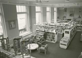 Picture of interior view of library branch showing shelves and chairs and people standing and s…