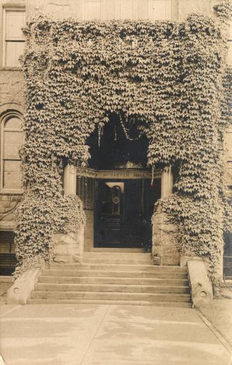 Black and white photograph of the ivy covered entrance to a very large late Victorian design bu…