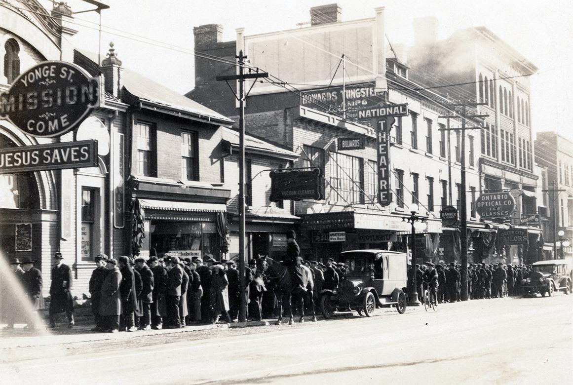 A photograph of a religious mission, with a long line of people on the sidewalk outside leading…