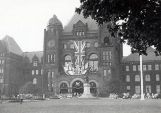 A photograph of the main entrance to a parliament building, decorated with bunting and flags of…