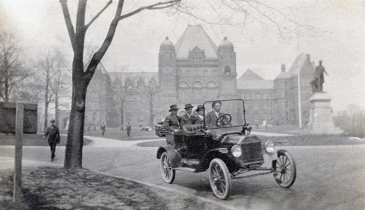 A photograph of four people in a car on a paved road with a large government building in the ba…