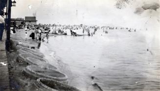 A photograph of a beach, with many people standing and sitting on the beach and swimming in the…