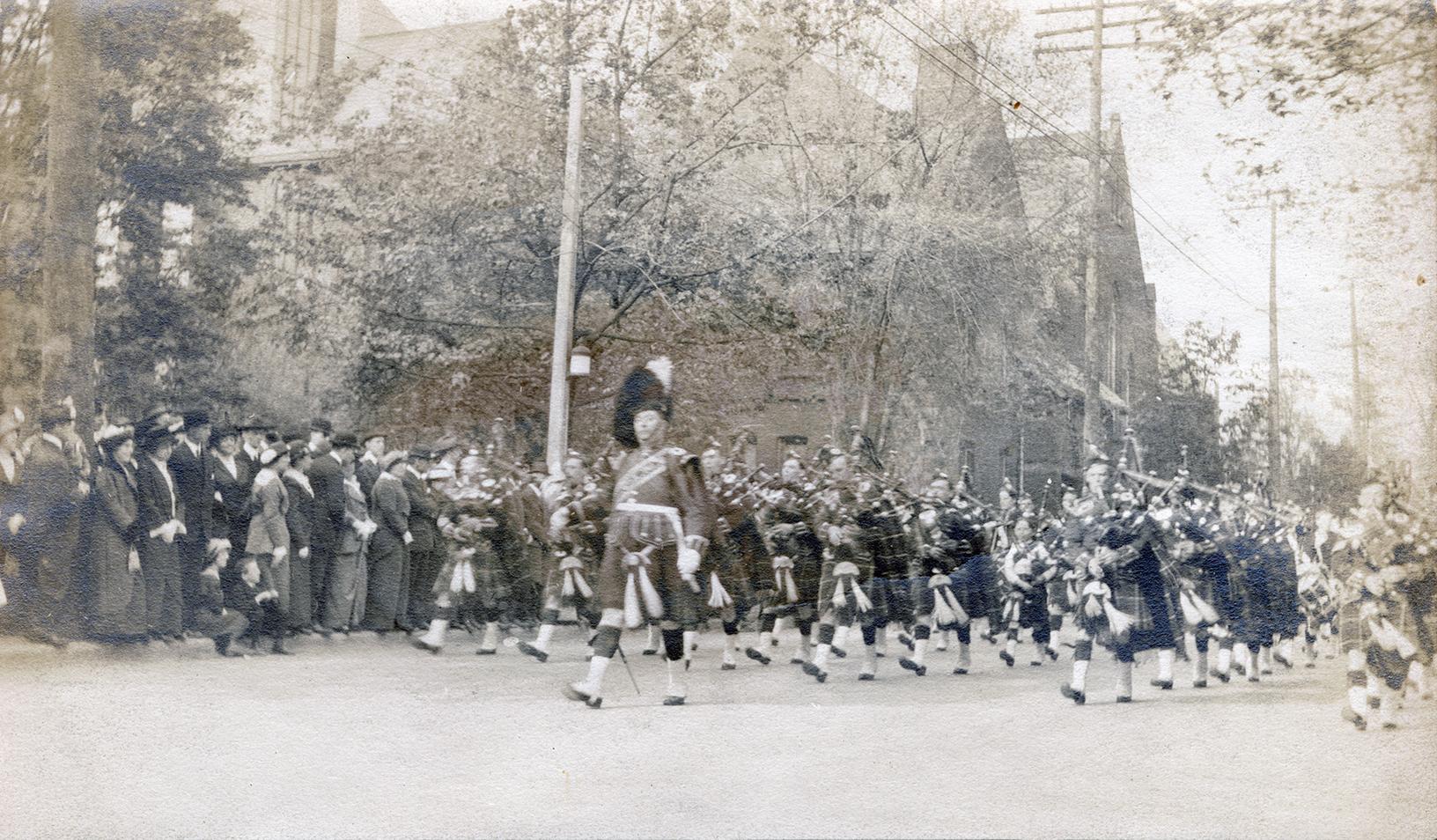 A photograph of a military parade, with dozens of men in kilts and hats carrying and playing ba…