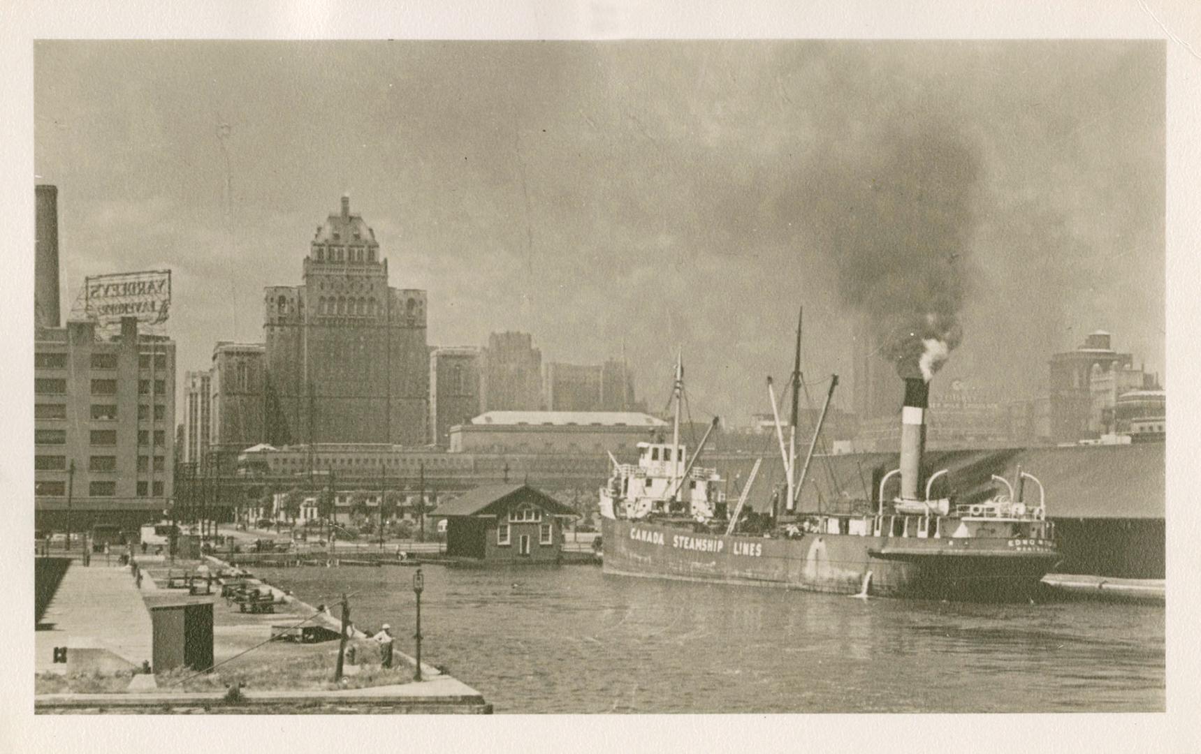 Black and white photo postcard depicting the Toronto harbourfront, dock, and steamship, with th…