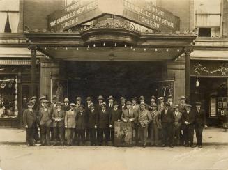 Black and white photograph of the Dumbells cast for "Cheerio" posing on the street in front of …