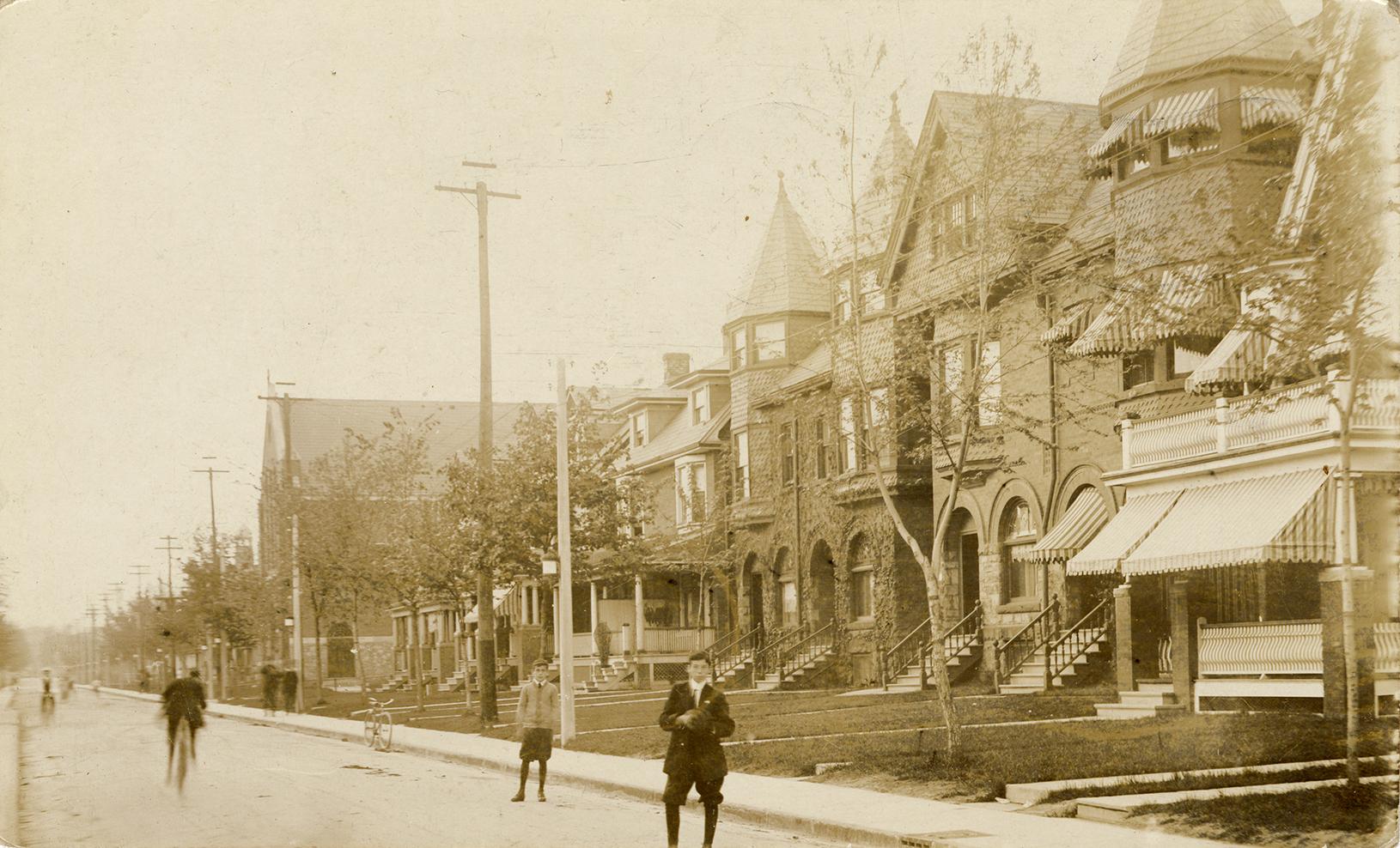 Black and white photograph of boys playing baseball on road with large houses on the side of it…