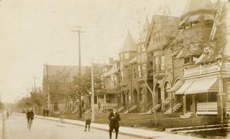 Black and white photograph of boys playing baseball on road with large houses on the side of it…