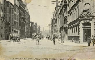 Black and white photo postcard depicting Wellington Street looking east from Yonge St. with bui…