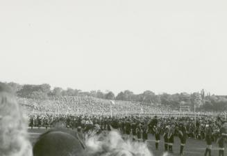 A photograph of a large crowd gathered in a park. There is a hill in the background and a flat …