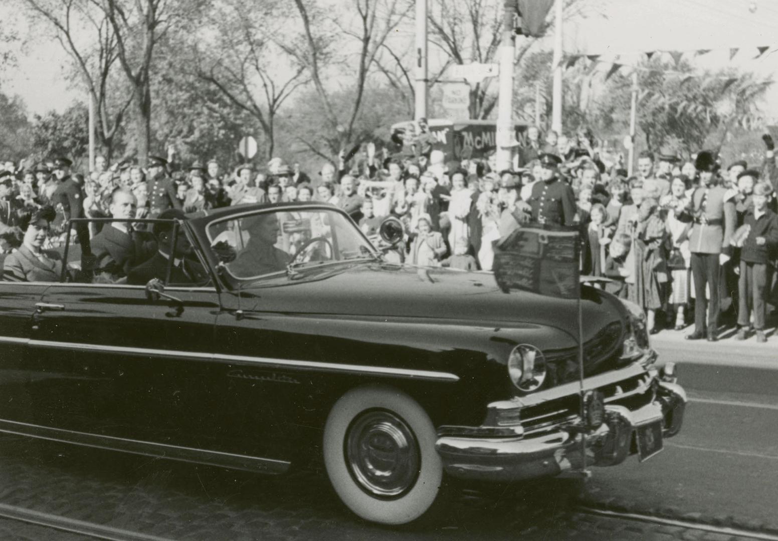 A photograph of car driving past a group of spectators on the sidewalk beside a paved city stre ...