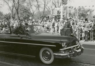 A photograph of car driving past a group of spectators on the sidewalk beside a paved city stre…