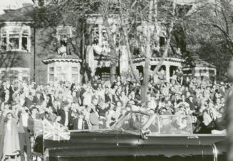 A photograph of a car driving past a crowd of spectators standing in front of a row of two stor ...