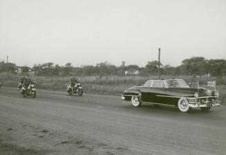 A photograph of a car driving down a paved road with a field and tress in the background. The c…