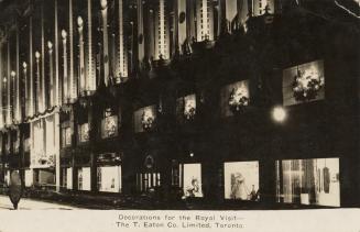 Black and white photograph of the front of a department store with banners at night with decora…