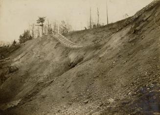 A photograph of a set of train tracks on top of and hanging down over the side of a dirt hill. …