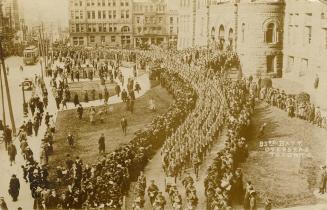 Black and white picture of soldiers marching in front of large, Richardsonian Romanesque buildi…