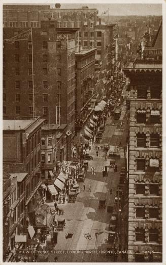 Black and white aerial photograph of skyscrapers on both sides of a busy street decorated with …