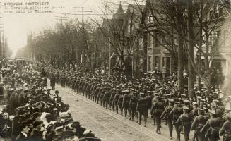 Black and white picture of soldiers marching on a city road with hundreds of civilian onlookers…