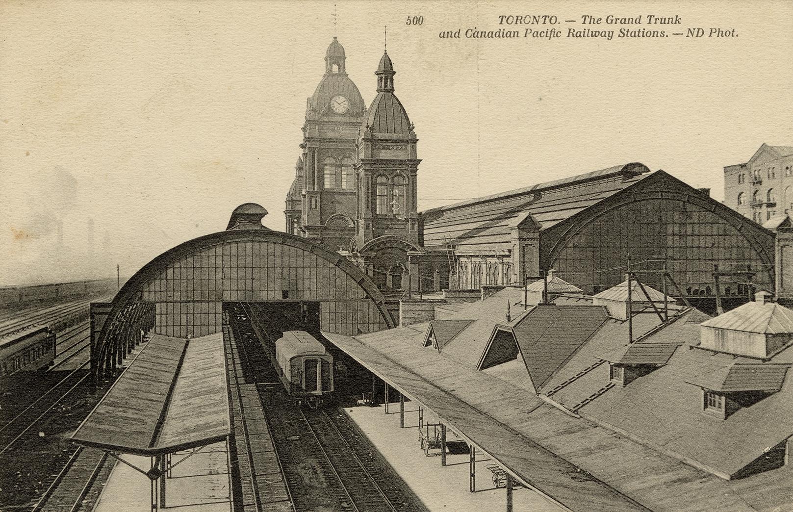 Black-white photo postcard depicting the exterior of a train station with the railroad yard, on…